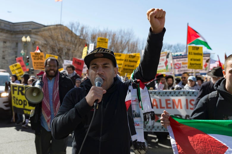 A demonstrator raises his fist and chants "free Palestine" at a protest. The crowd behind him marches with signs calling for an independent Palestinian state.